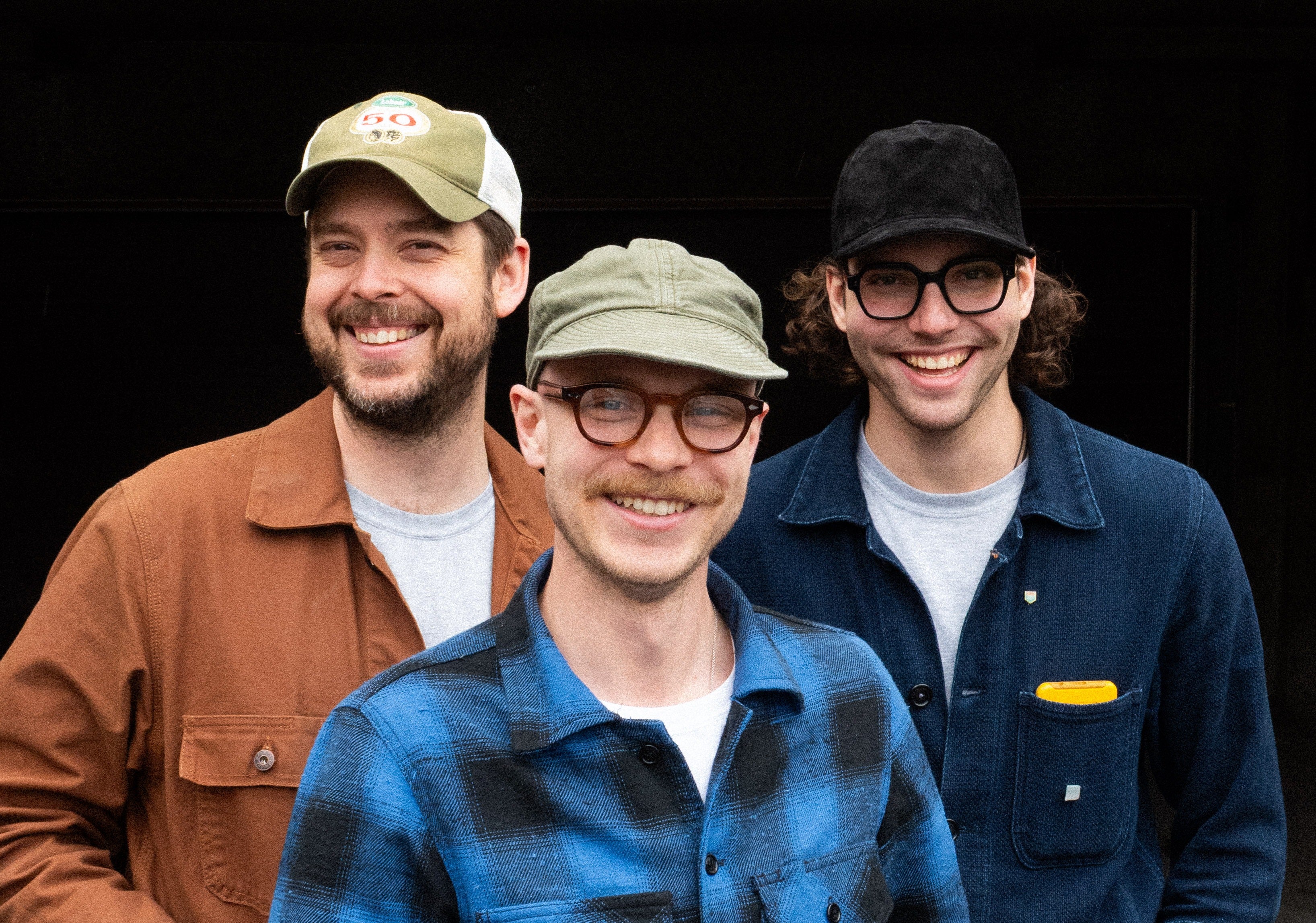 Three men wearing hats and smiling against a black background